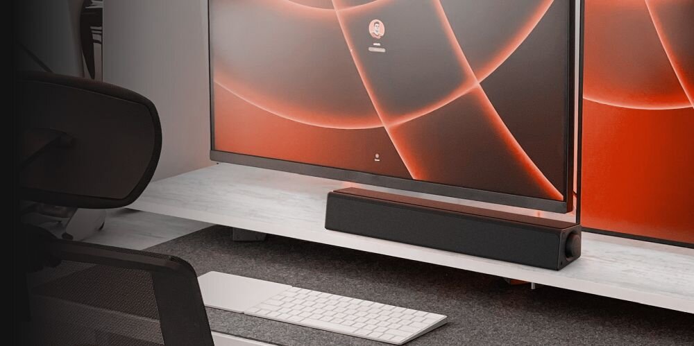 Na białej półce znajduje się czarny soundbar przed dużym monitorem z pomarańczowym wzorem na ekranie. Przed półką leży biała klawiatura i myszka.