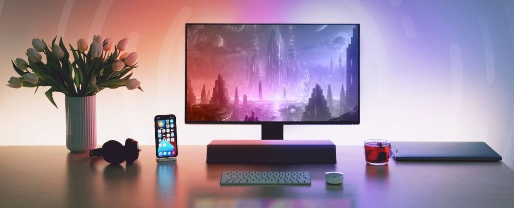 Na biurku znajduje się monitor z wyświetlanym krajobrazem futurystycznego miasta, obok którego stoi smartfon, klawiatura, myszka, filiżanka z napojem oraz zamknięty laptop. Obok monitora widoczny jest wazon z kwiatami.