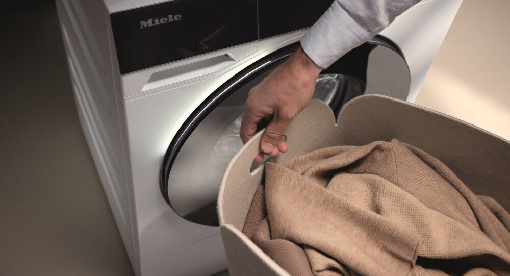 Osoba wkłada pranie do bębna pralki marki Miele. Na pralce widoczne jest logo \'Miele\'.