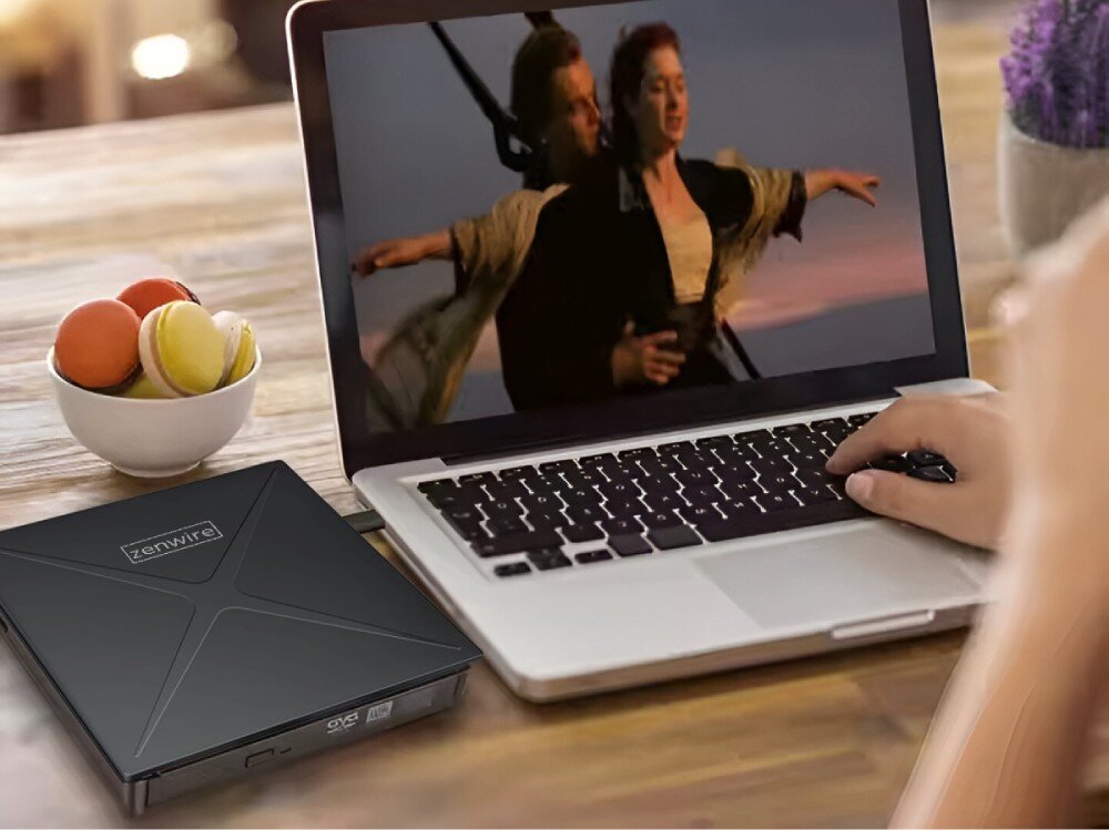 Na stole znajduje się otwarty laptop z wyświetlanym filmem oraz czarny napęd z logo \'zenwire\'. Obok laptopa stoi miska z kolorowymi makaronikami.