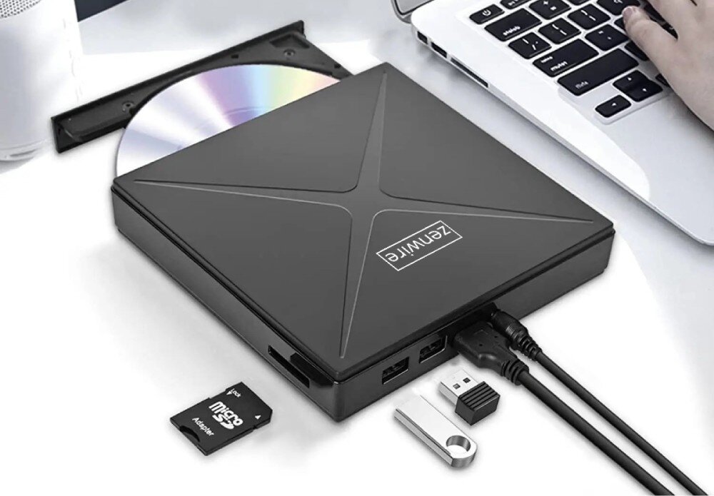 Na obrazie znajduje się czarny napęd z logo \'ZENWIRE\', z otwartą tacką na płytę CD. Obok napędu leżą różne akcesoria, w tym karta microSD, pendrive i adapter USB, a w tle widoczna jest klawiatura laptopa.