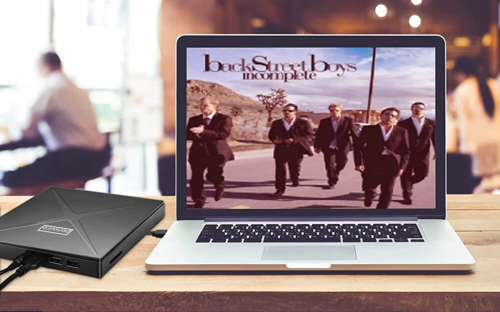 Na drewnianym stole stoi otwarty laptop, na ekranie którego widoczna jest okładka albumu z napisem \'Backstreet Boys incomplete\'. Obok laptopa znajduje się czarny napęd z logo \'ZENWIRE\'.