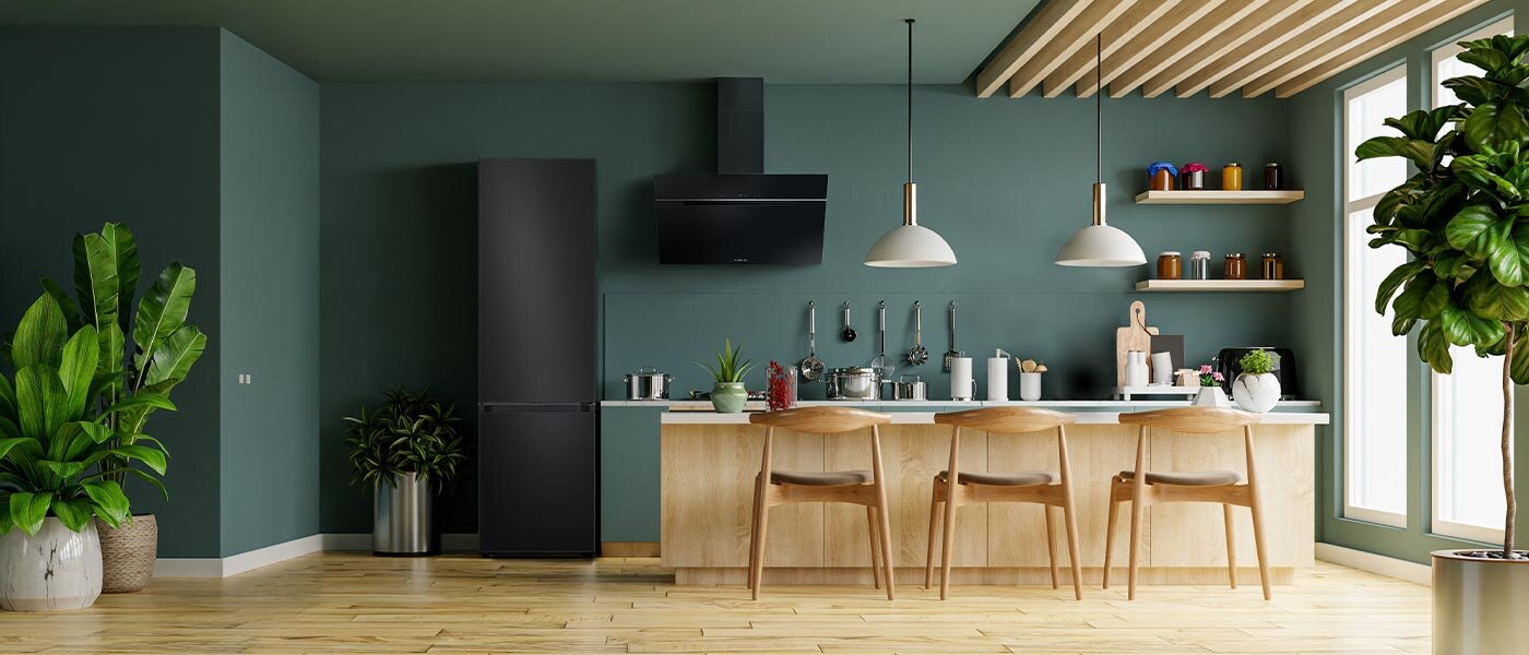 Lodówka z dolnym zamrażalnikiem Samsung RB38C7B6AB1 z oferty Media Expert wstawiona do ciemnozielonego pomieszczenia kuchennego Kuchnia z ciemnozielonymi ścianami, w której znajduje się czarna lodówka obok drewnianego blatu z czterema krzesłami. Na blacie stoją różne akcesoria kuchenne, a nad nim wiszą dwie lampy.