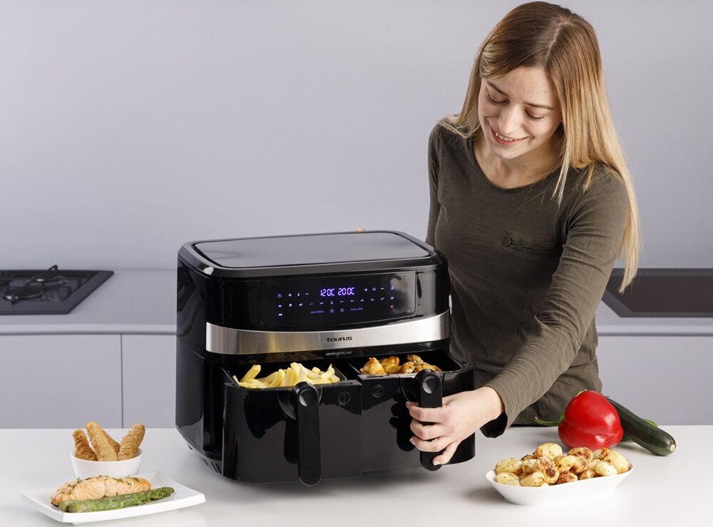 Kobieta obsługuje czarną frytkownicę beztłuszczową z podwójnym koszem, na której wyświetlaczu widnieje \'200°C 20:00\'. Obok frytkownicy znajdują się talerze z jedzeniem oraz papryka i cukinia.
