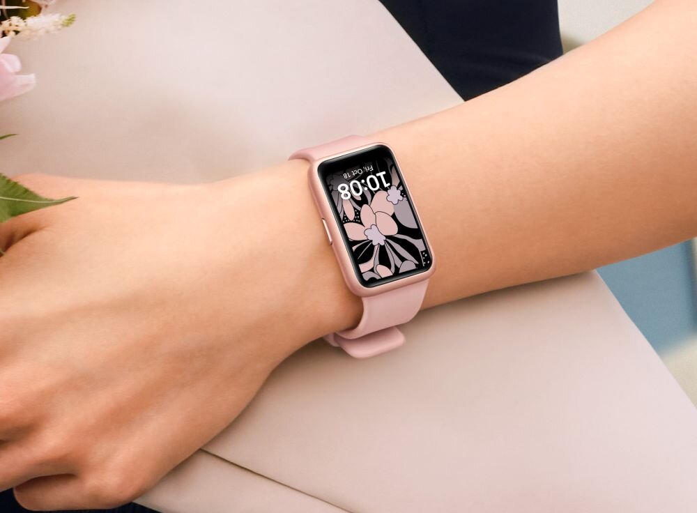 Na zdjęciu widoczny jest różowy smartwatch na nadgarstku, z wyświetlaczem pokazującym godzinę 10:08 i grafiką kwiatową. Pasek zegarka jest również różowy.