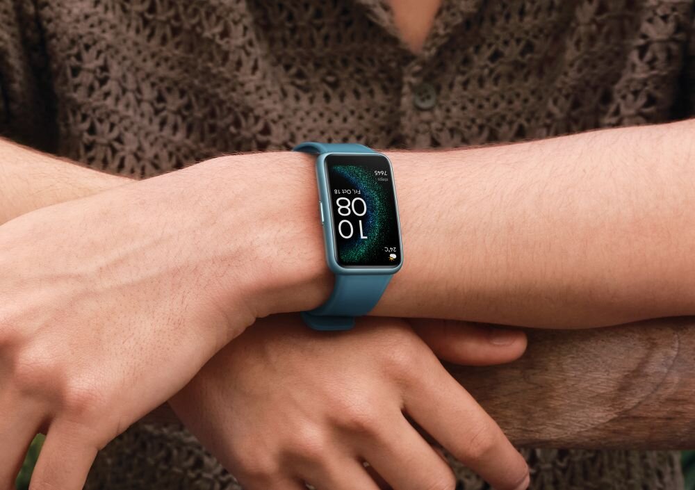 Osoba nosi smartwatch z prostokątnym wyświetlaczem, na którym widoczna jest godzina 10:08. Pasek zegarka jest w kolorze niebieskim.
