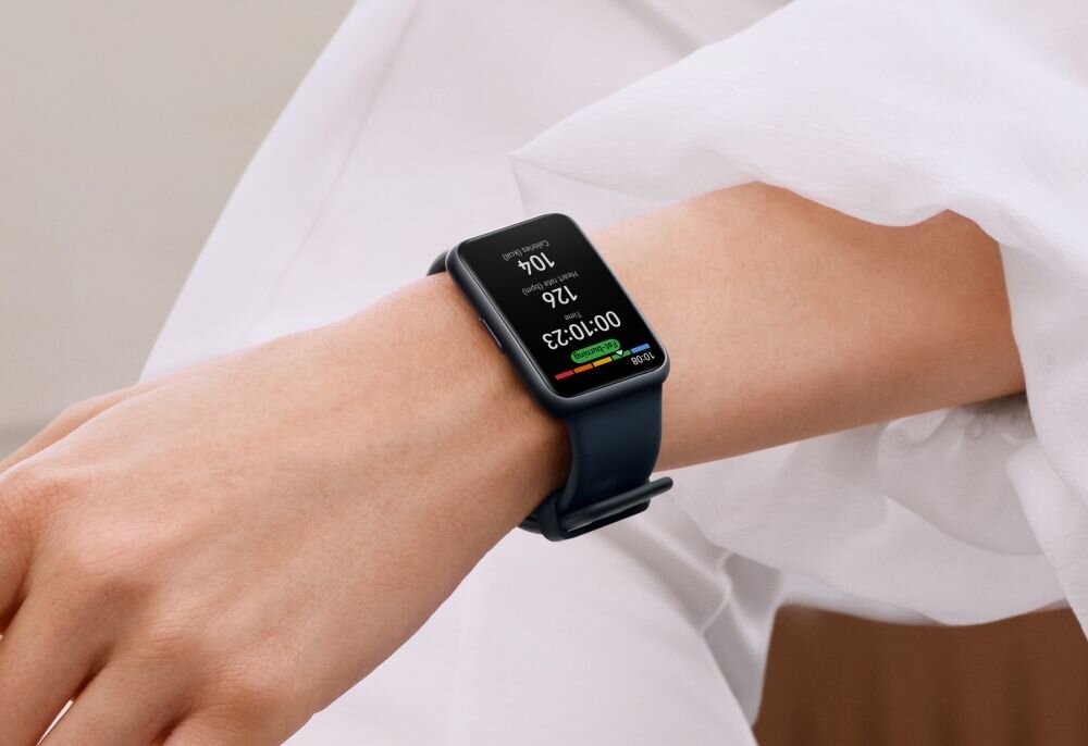 Osoba nosi smartwatch z prostokątnym wyświetlaczem, na którym widoczny jest tekst: \'00:10:23\', \'126\', \'10k\', \'Calories burned\'. Pasek zegarka jest ciemny, a tło stanowi jasna odzież.
