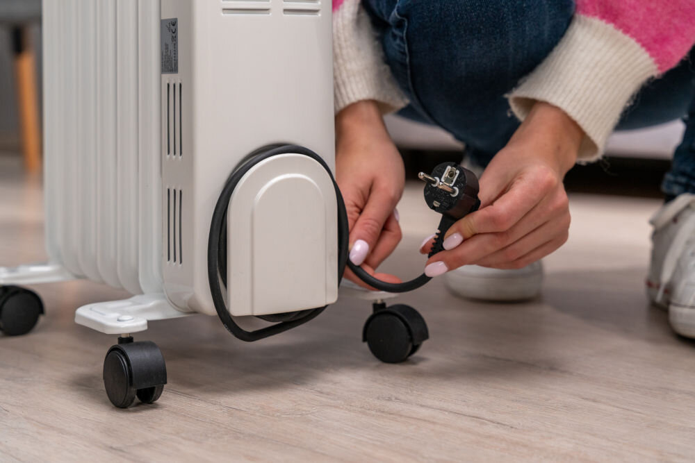 Osoba trzyma wtyczkę elektryczną przy grzejniku olejowym na kółkach, stojącym na podłodze.
