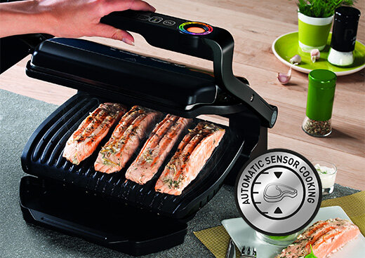 Na obrazie widoczny jest grill elektryczny z kilkoma kawałkami łososia na ruszcie. Obok znajduje się okrągły symbol z napisem \'AUTOMATIC SENSOR COOKING\'.