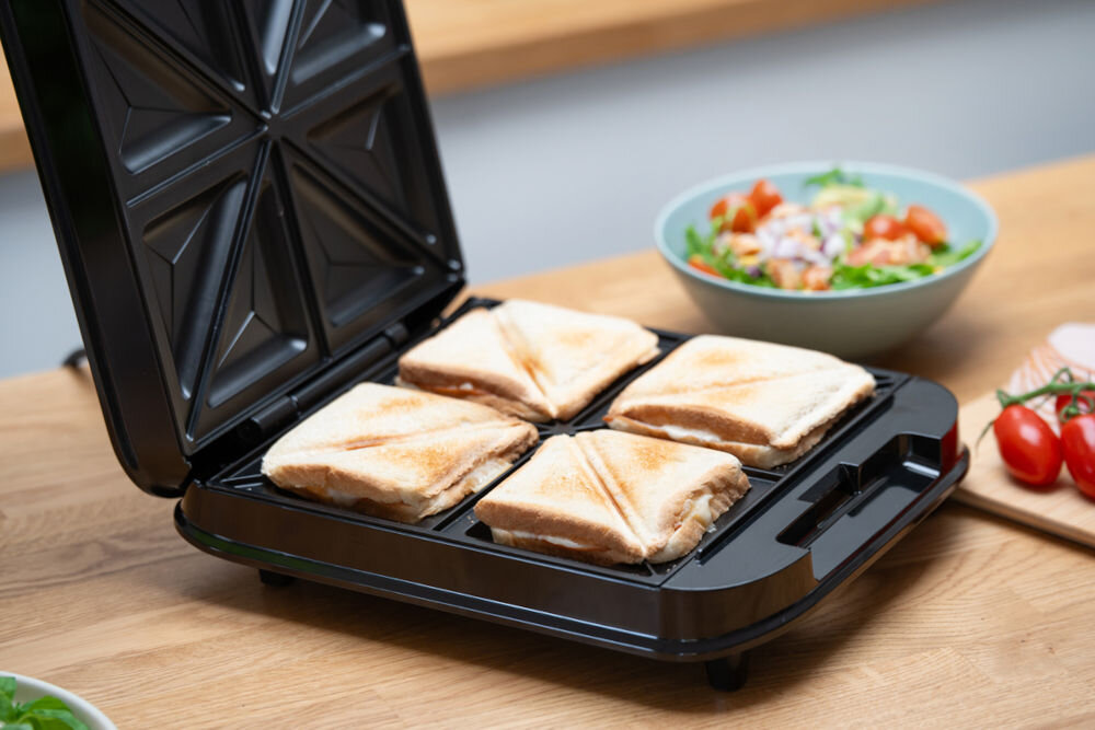 Na drewnianym blacie stoi otwarty opiekacz z czterema tostami. W tle widoczne są miska z sałatką i kilka pomidorów koktajlowych.