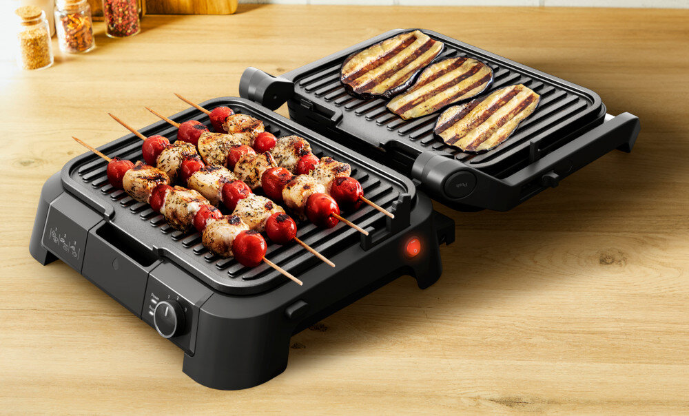 Na drewnianym blacie znajduje się otwarty grill elektryczny z dwoma powierzchniami grillującymi. Na jednej części leżą szaszłyki z mięsem i pomidorami, a na drugiej plastry bakłażana.