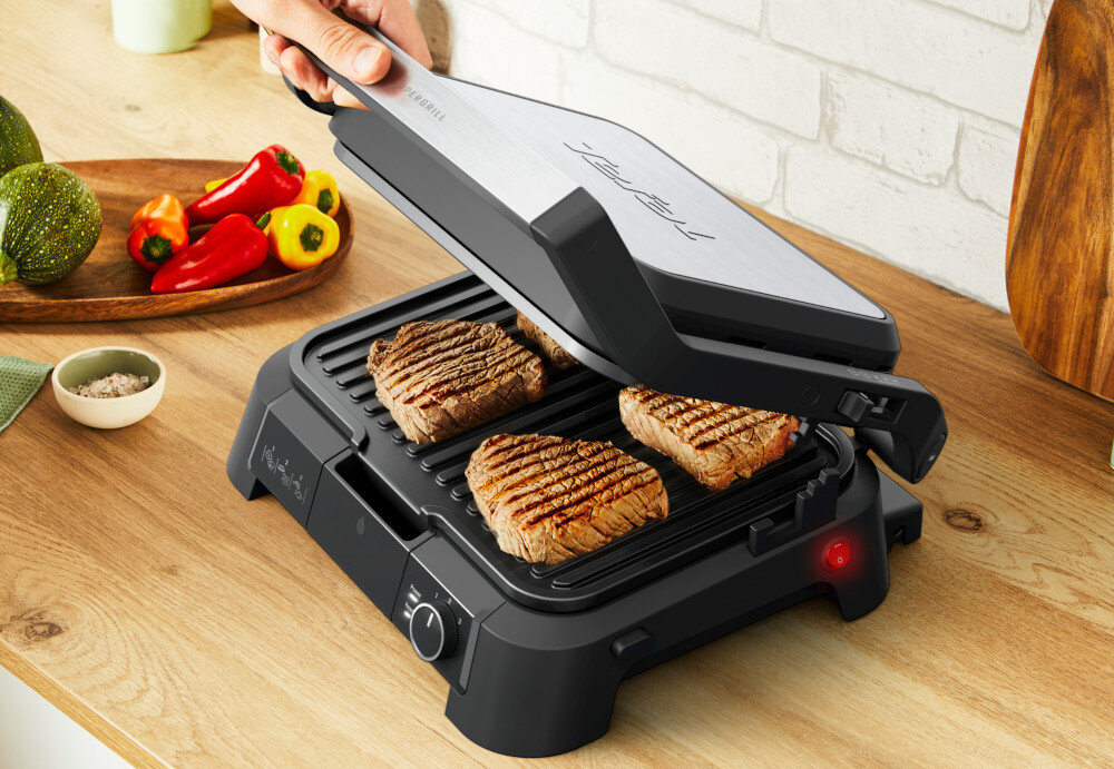Na drewnianym blacie znajduje się otwarty grill elektryczny z trzema kawałkami mięsa. Obok leży talerz z kolorowymi paprykami i cukinią.