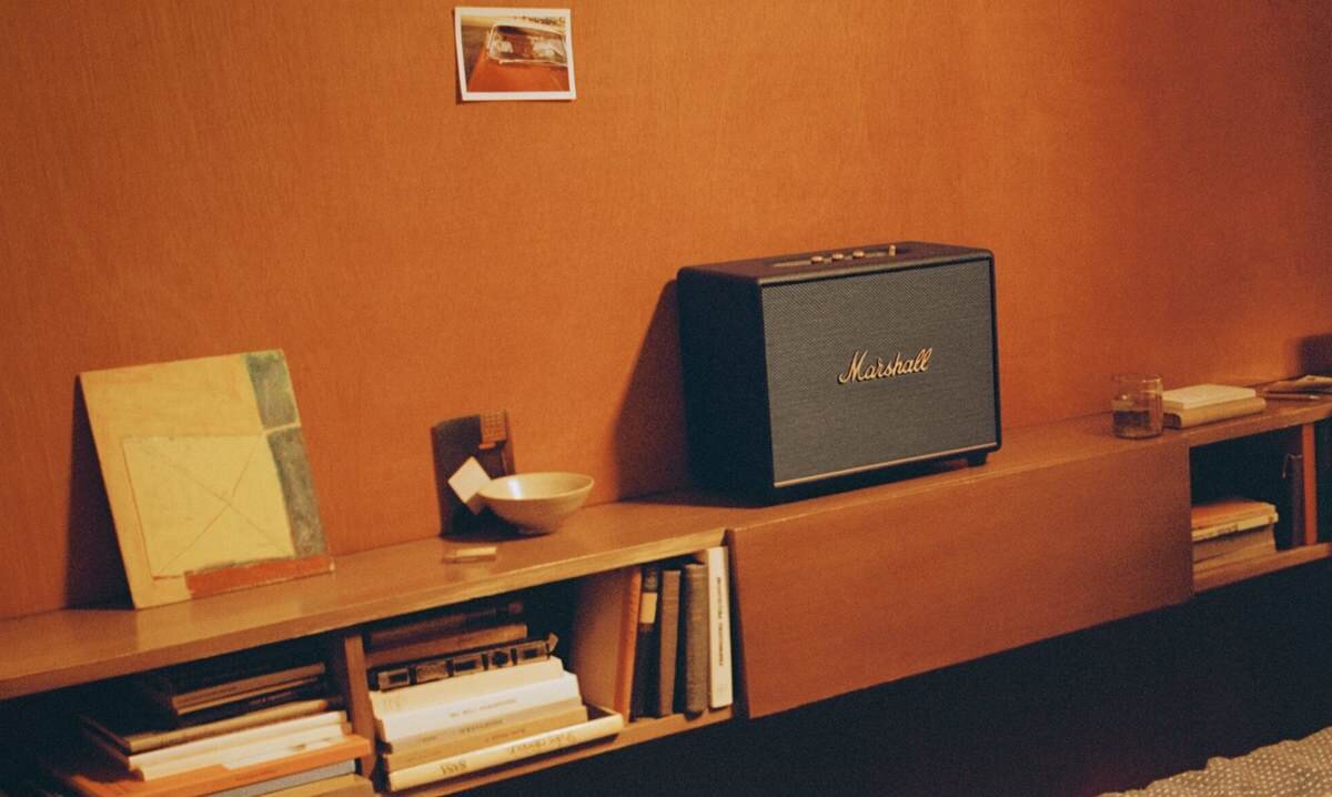 Na drewnianej półce znajduje się czarny głośnik z logo \'Marshall\'. Obok głośnika leżą książki i inne drobne przedmioty.