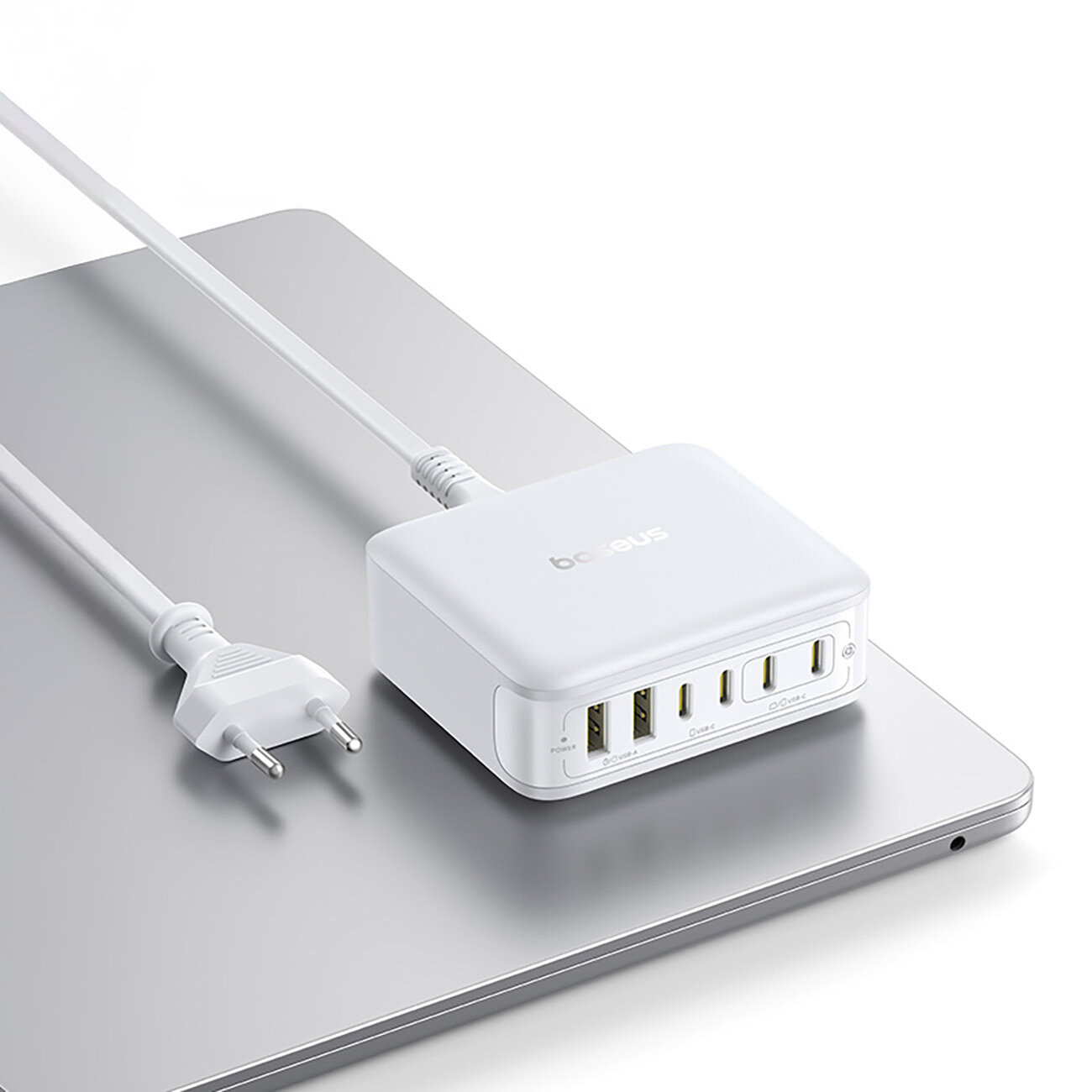 Na obrazie widoczna jest biała stacja ładująca z logo \'baseus\', posiadająca cztery porty USB-C i dwa porty USB-A. Urządzenie leży na srebrnym laptopie, a do stacji podłączony jest biały kabel z wtyczką.