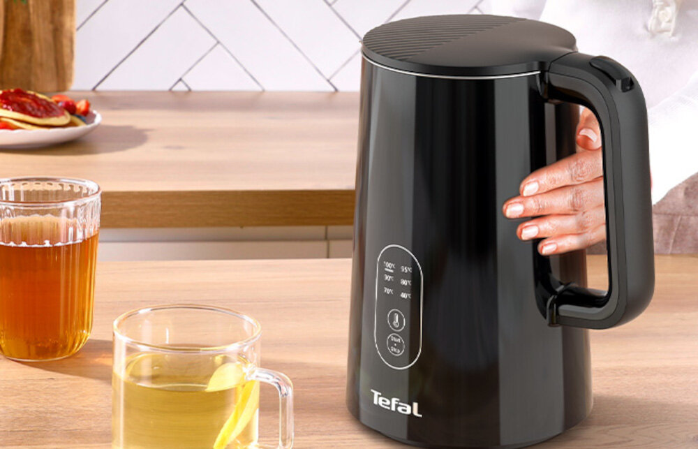 Czarny czajnik elektryczny z logo Tefal i przyciskami do regulacji temperatury stoi na blacie kuchennym. Obok znajdują się dwie szklanki z napojem, a w tle widać talerz z jedzeniem.