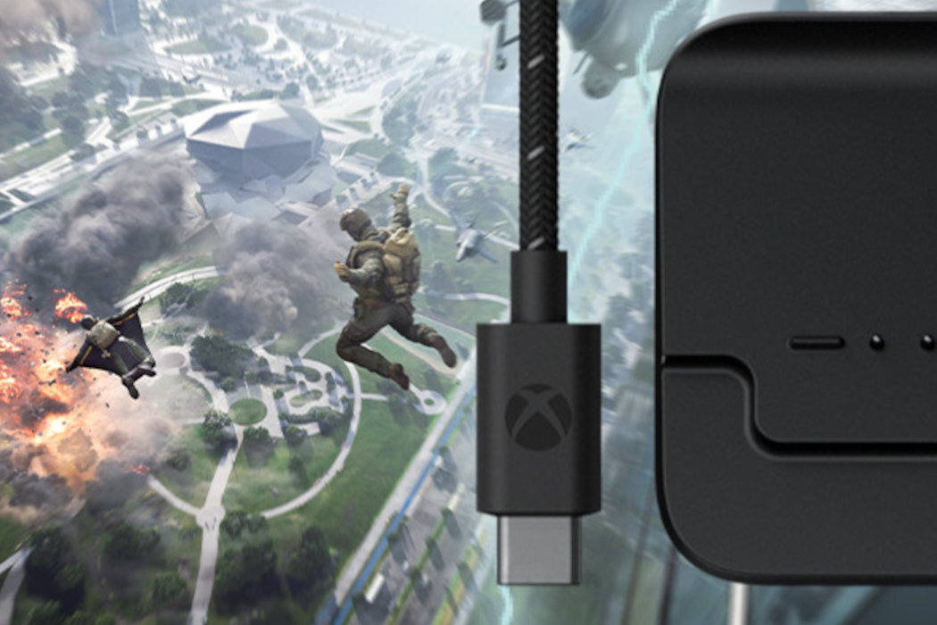 Na obrazie widoczny jest czarny kabel z logo Xbox oraz fragment czarnego urządzenia. W tle znajduje się dynamiczna scena z dwoma postaciami w powietrzu i eksplozją na ziemi.