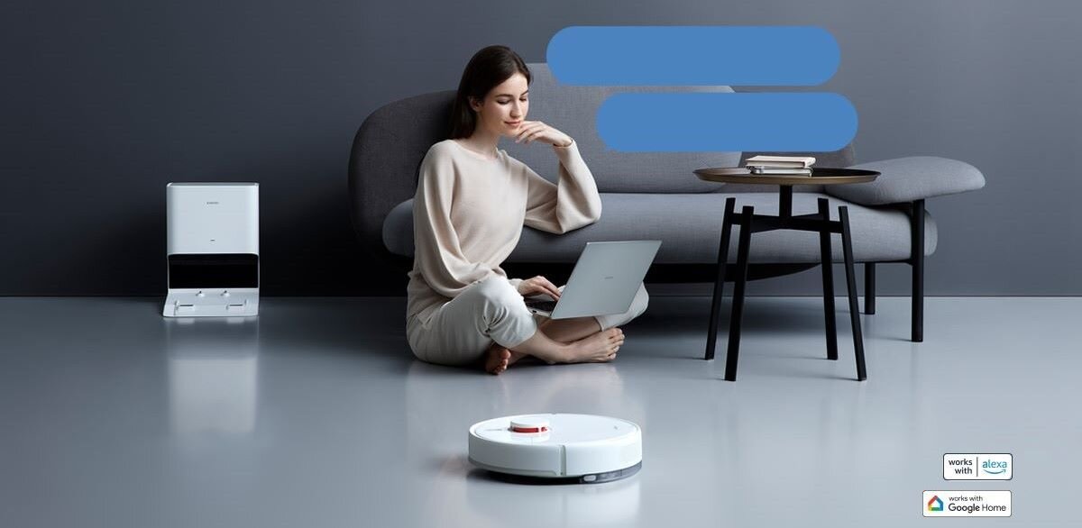 Kobieta siedzi na podłodze z laptopem, obok niej znajduje się biały robot sprzątający. W tle widać sofę, stolik z książkami oraz stację dokującą.