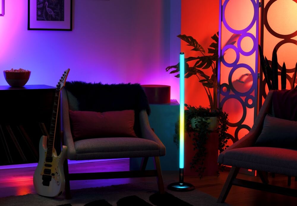 W pomieszczeniu znajduje się kolorowa lampa stojąca emitująca światło w różnych barwach. Obok lampy widoczne są dwa fotele, gitara oraz dekoracyjna ścianka z okręgami.
