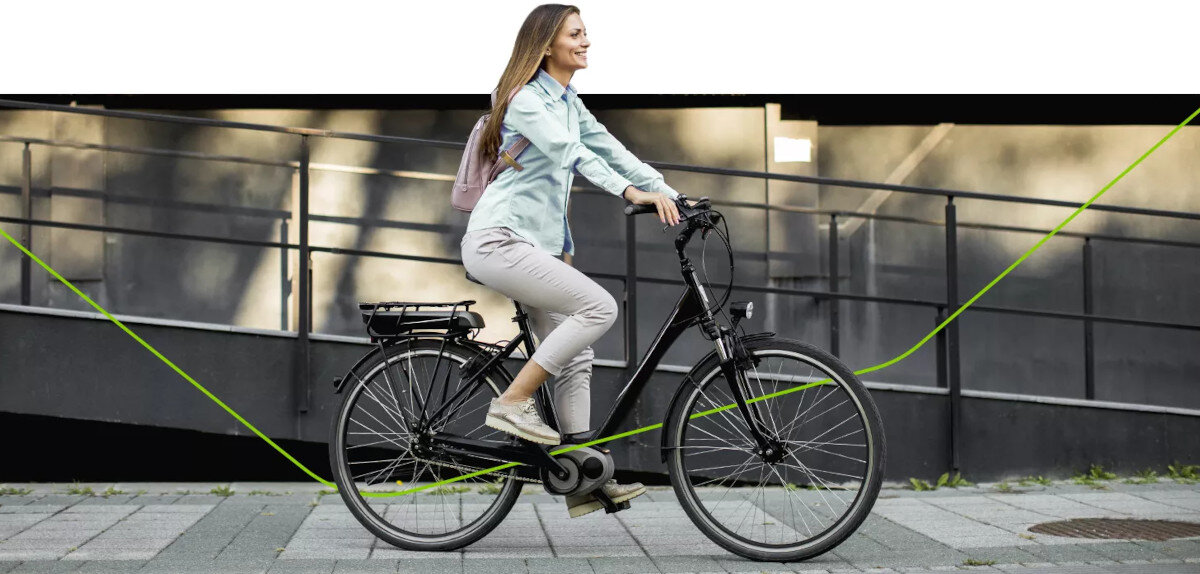 Osoba jedzie na czarnym rowerze elektrycznym po chodniku, w tle widoczna jest metalowa barierka. Rower wyposażony jest w baterię zamontowaną na bagażniku.