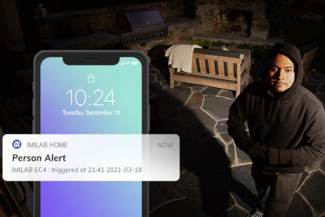 Na obrazie widać ekran telefonu z powiadomieniem \'Person Alert\' od aplikacji IMILAB HOME, z datą i godziną \'triggered at 21:41 2021-03-18\'. W tle znajduje się osoba w ciemnym ubraniu na kamiennej posadzce, obok drewnianej ławki i grilla.