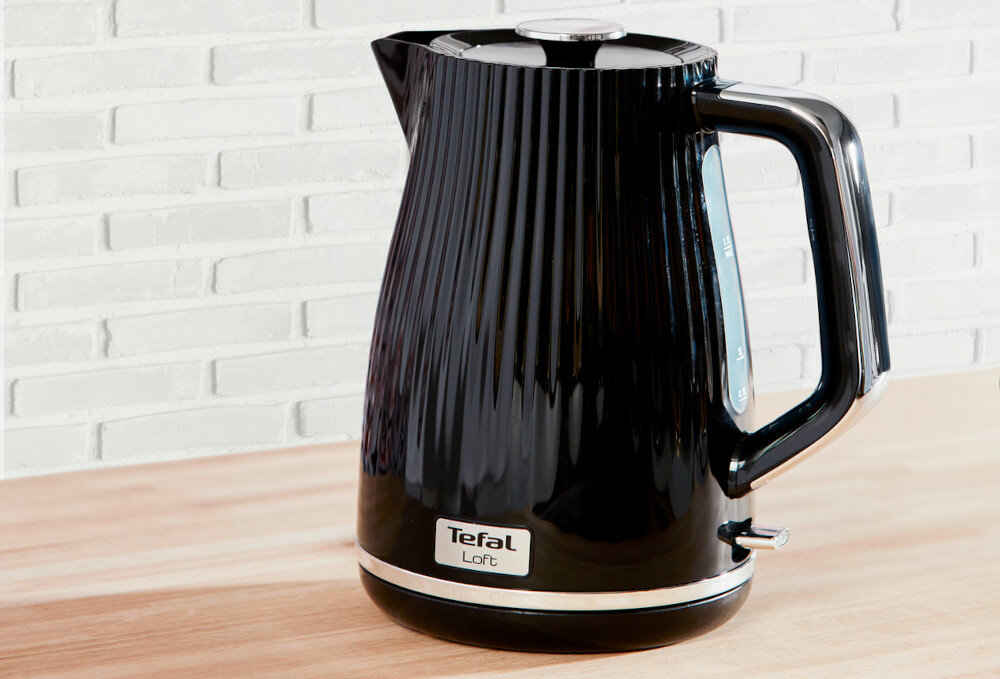 Czarny czajnik elektryczny z pionowymi żłobieniami, z logo \'Tefal Loft\' na dole. Stoi na drewnianym blacie, w tle widoczna jest biała ceglana ściana.