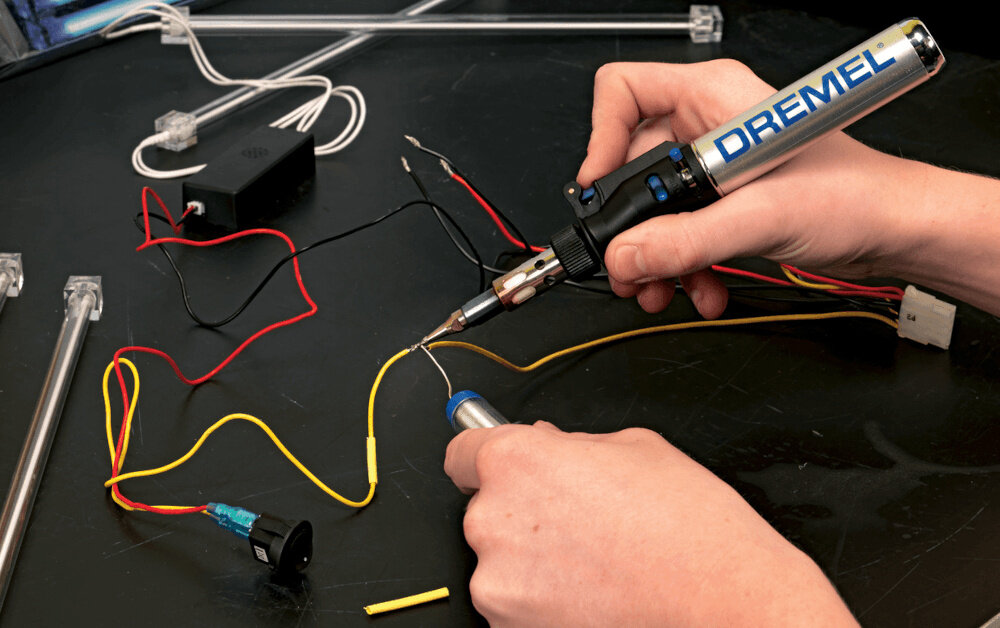 Osoba używa lutownicy z widocznym logo \'DREMEL\' do łączenia przewodów elektrycznych na czarnym stole. W tle widoczne są różne przewody i elementy elektroniczne.