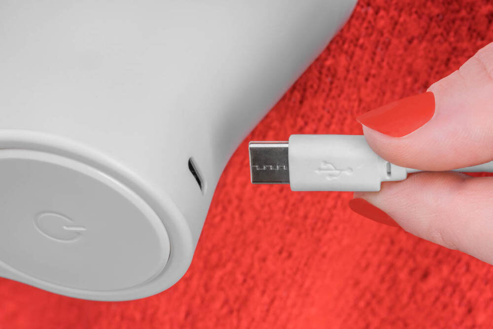 Osoba trzyma wtyczkę USB, zbliżając ją do portu w urządzeniu o gładkiej, jasnej powierzchni. W tle widoczny jest czerwony materiał, a na urządzeniu znajduje się litera \'G\'.