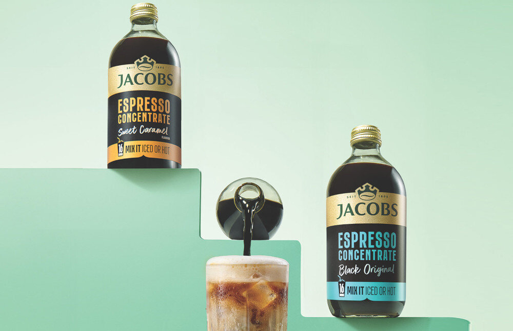Na obrazie znajdują się dwie butelki koncentratu kawowego marki JACOBS, jedna z napisem \'Espresso Concentrate Sweet Caramel\', a druga \'Espresso Concentrate Black Original\'. Obok butelek widoczna jest szklanka z kawą z lodem.