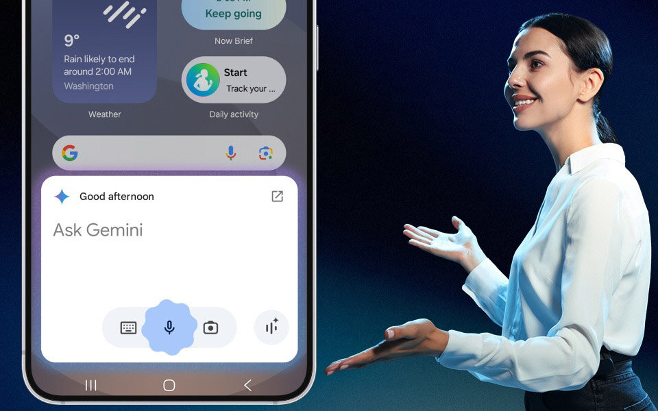 Obraz przedstawia smartfon z wyświetlaczem pokazującym aplikację z tekstem \'Good afternoon\' i \'Ask Gemini\'. Obok stoi osoba w białej koszuli, gestykulująca rękami.