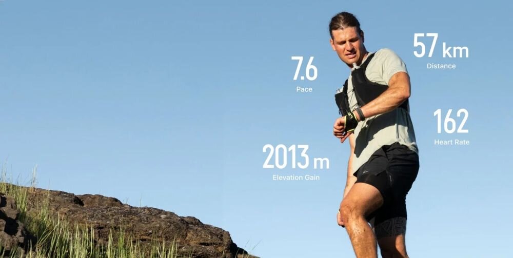 Osoba biegnie po skalistym terenie, a na niebie widoczne są dane: 7.6 Pace, 57 km Distance, 162 Heart Rate, 2013 m Elevation Gain. Osoba ma na ręce smartwatch i jest ubrana w sportowy strój.