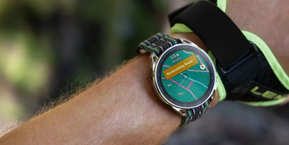 Osoba nosi smartwatch z okrągłym wyświetlaczem, na którym widoczna jest mapa z napisem \'Recalculating Route\'. Pasek zegarka ma wzór w ciemnych kolorach.