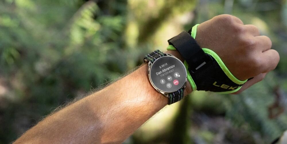 Osoba nosi smartwatch na nadgarstku, na ekranie widoczny jest przychodzący telefon od \'Dan Abramov\'. W tle znajduje się rozmyta zieleń, sugerująca otoczenie przyrody.