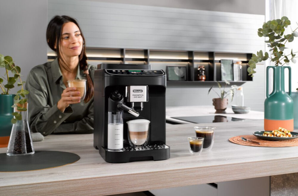 Na blacie kuchennym stoi czarny ekspres do kawy z logo \'DELONGHI\'. Obok ekspresu znajduje się filiżanka z kawą, a w tle widać osobę trzymającą szklankę z napojem.