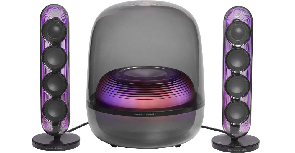 Na obrazie widoczny jest zestaw głośników z przezroczystymi obudowami, w tym subwoofer i dwa pionowe głośniki satelitarne. Na subwooferze znajduje się logo \'harman/kardon\'.