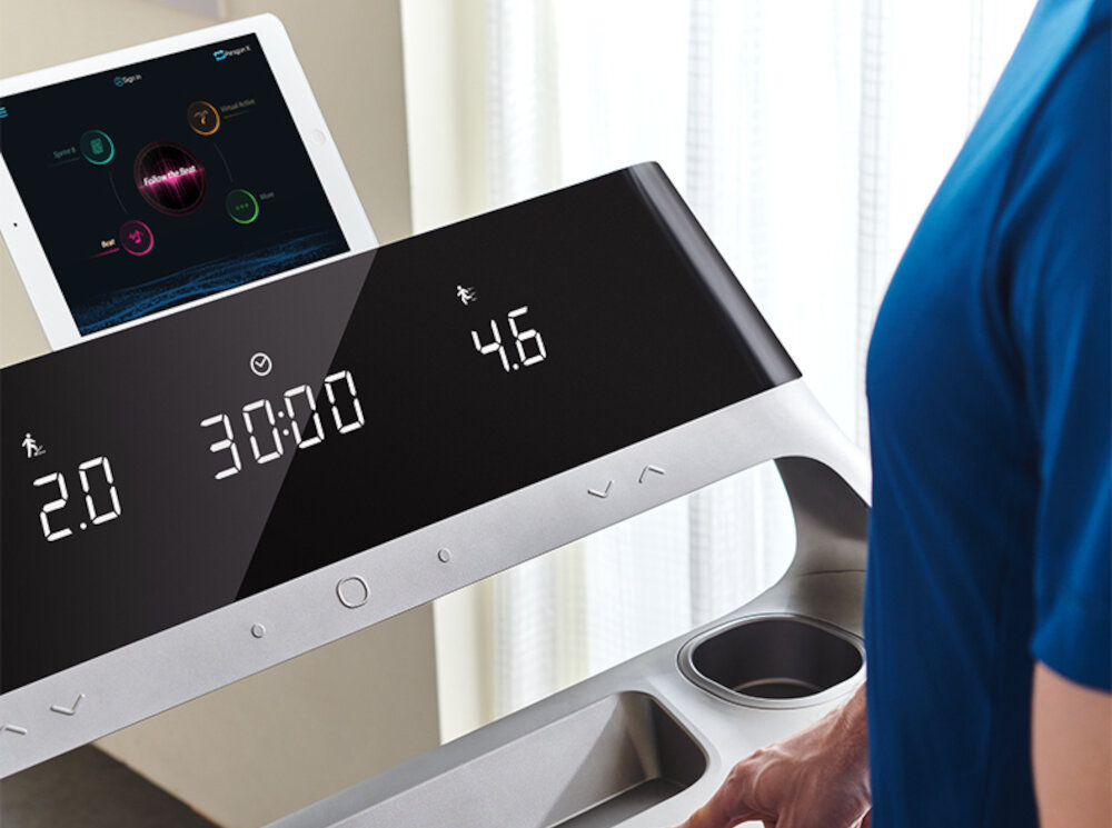 Panel sterowania bieżni elektrycznej z wyświetlaczem pokazującym wartości: 2.0, 30:00, 4.6. Na bieżni znajduje się tablet z aplikacją fitness.