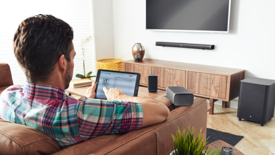 Osoba siedzi na kanapie, trzymając tablet, przed nią znajduje się telewizor z zamontowanym soundbarem. Na szafce pod telewizorem stoi głośnik, a obok znajduje się subwoofer.