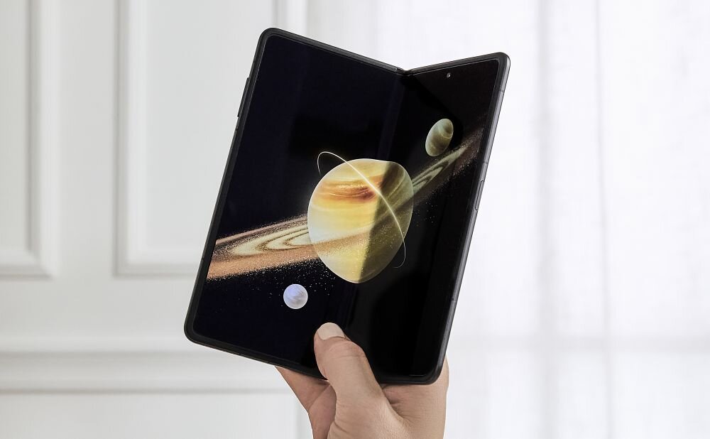 Osoba trzyma składany smartfon, na którego ekranie widoczna jest grafika przedstawiająca planety. W tle znajduje się jasna ściana z oknem.