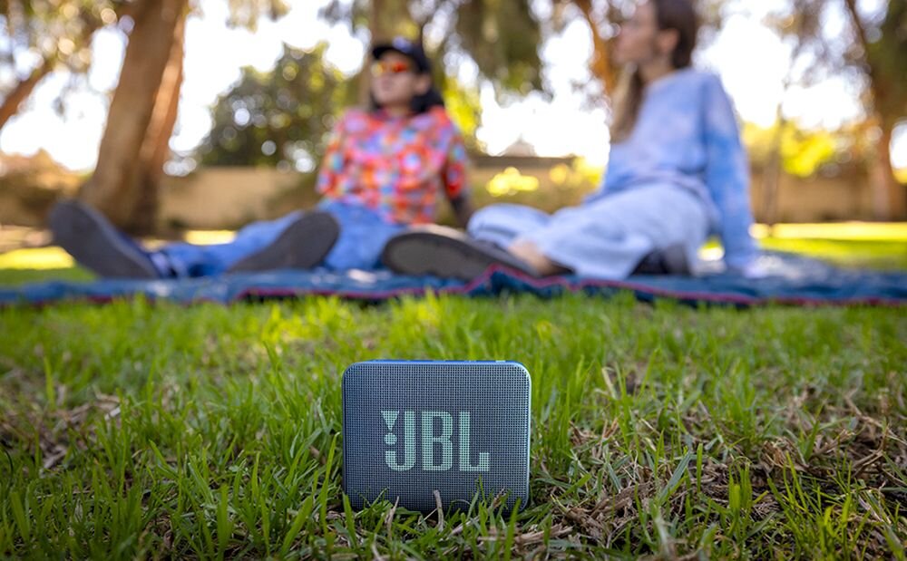 Na trawie znajduje się czarny głośnik z widocznym logo \'JBL\'. W tle dwie osoby siedzą na kocu w parku.