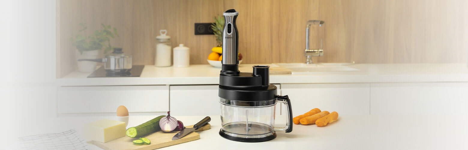 Na blacie kuchennym stoi blender z przezroczystym pojemnikiem i czarnymi elementami. W tle widoczne są warzywa, nóż oraz inne akcesoria kuchenne.