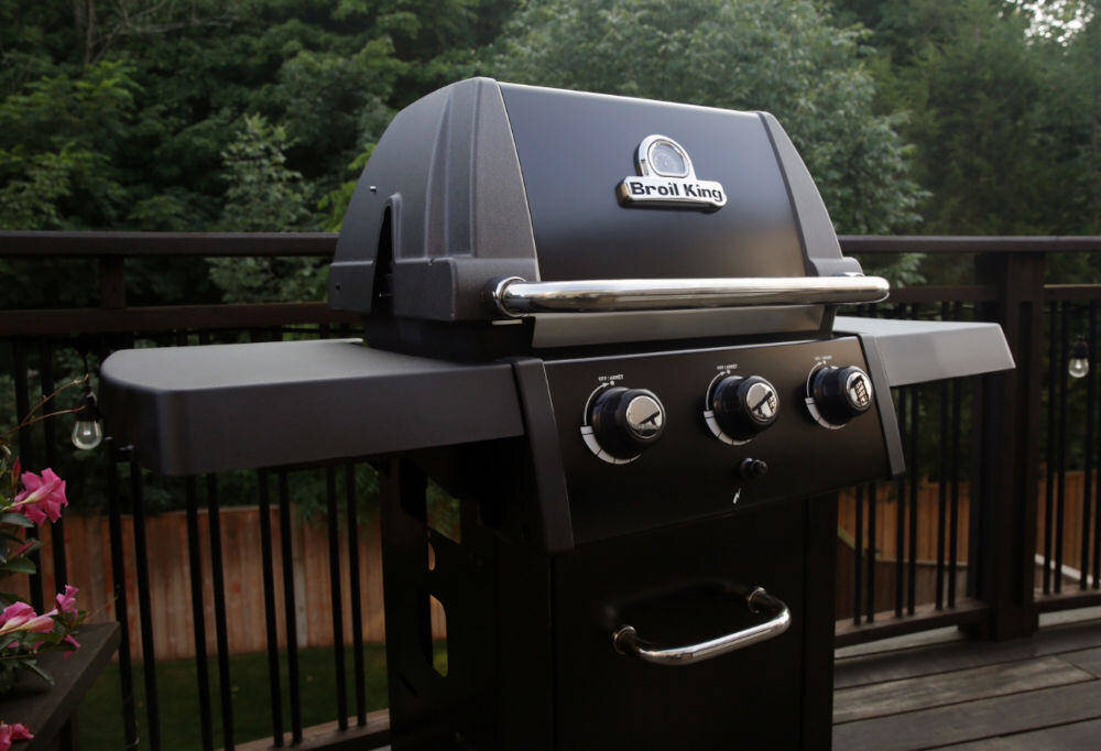 Na obrazie widoczny jest grill ogrodowy z trzema pokrętłami i logo \'Broil King\' na pokrywie. W tle znajdują się drzewa i drewniane ogrodzenie.