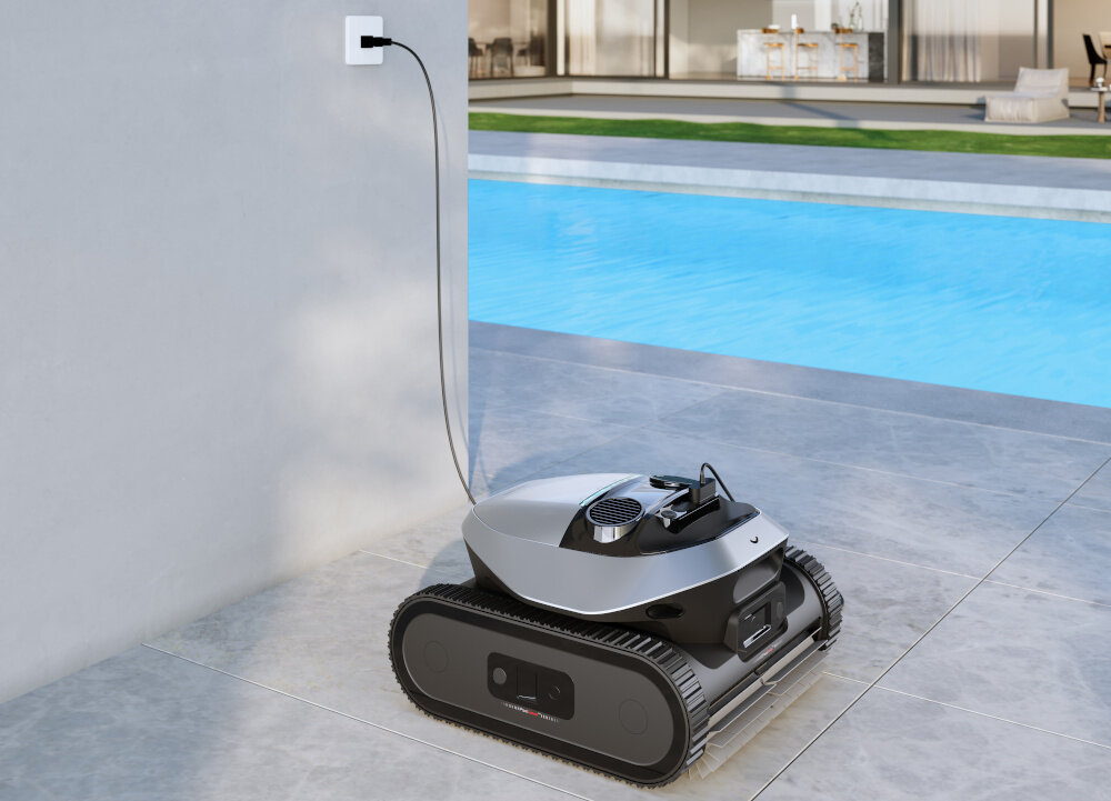 Na obrazie widoczny jest robot do czyszczenia basenów stojący na płytkach przy basenie, podłączony do gniazdka elektrycznego. W tle widać wodę w basenie oraz część budynku z dużymi oknami.
