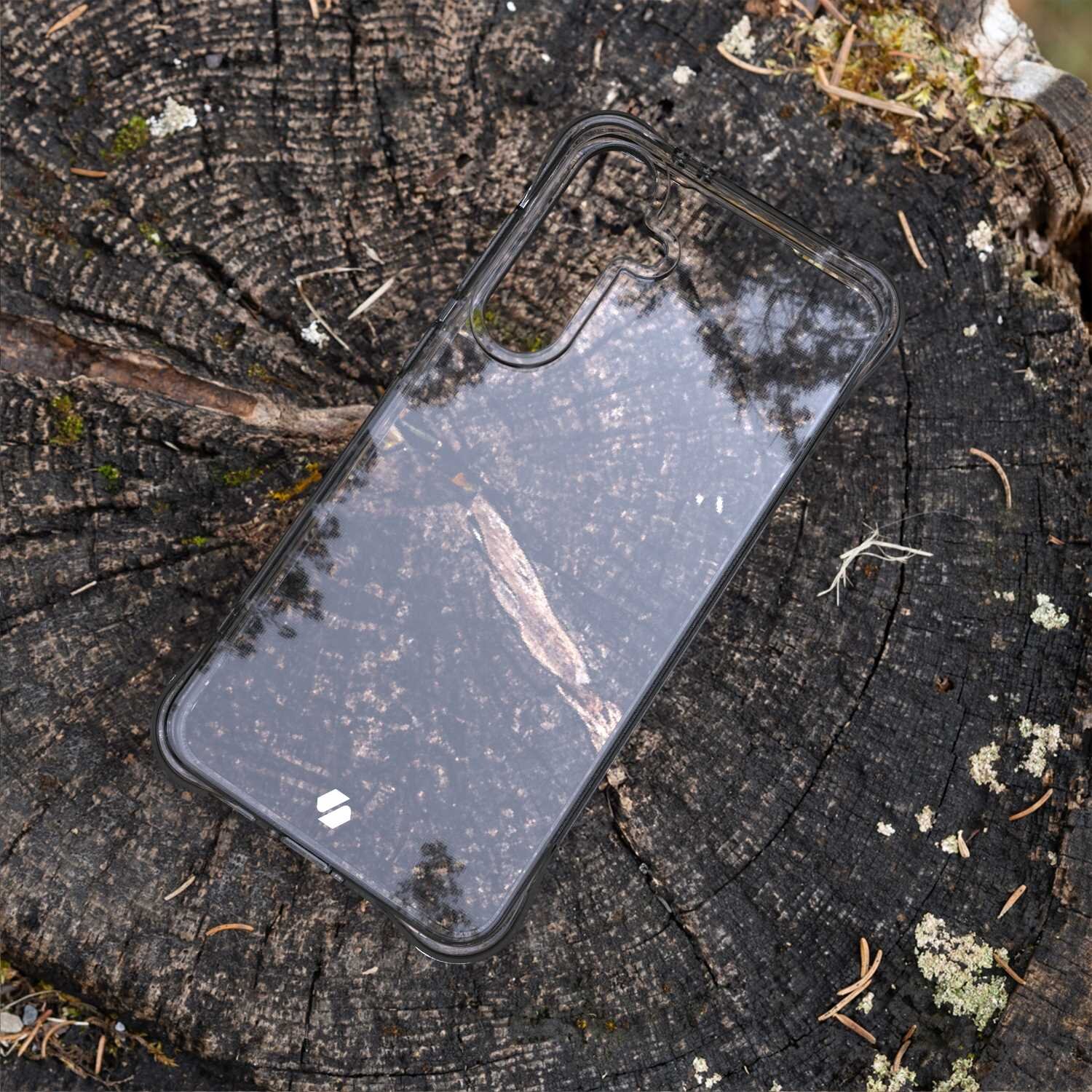 Przezroczyste silikonowe etui na telefon leży na pniu drzewa, ukazując jego strukturę i słoje. Na etui widoczne jest małe logo producenta.