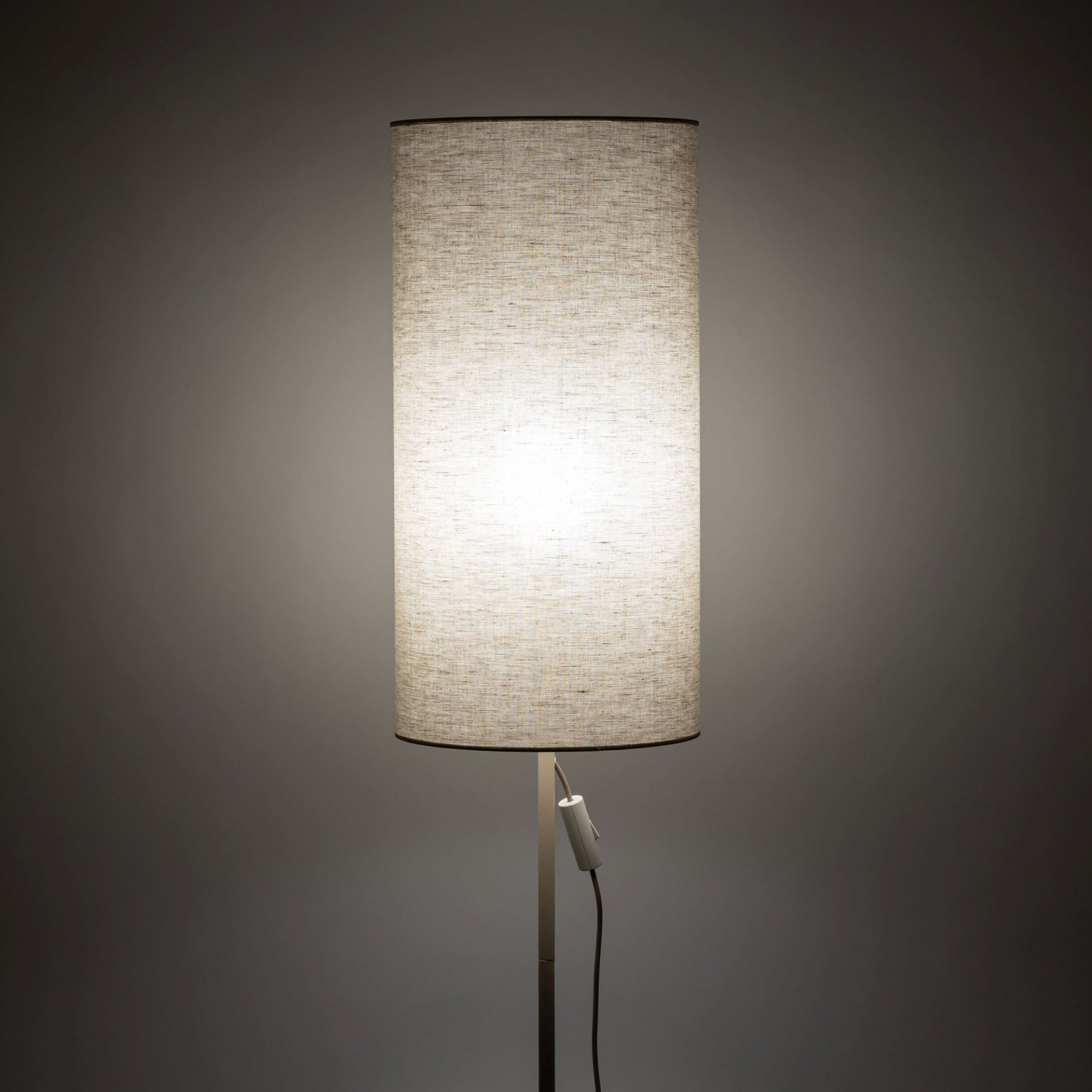 Na obrazie widoczna jest lampa podłogowa z beżowym, cylindrycznym abażurem wykonanym z materiału przypominającego len. Lampa jest włączona, co powoduje, że abażur jest równomiernie oświetlony, a na stelażu znajduje się włącznik z przewodem.