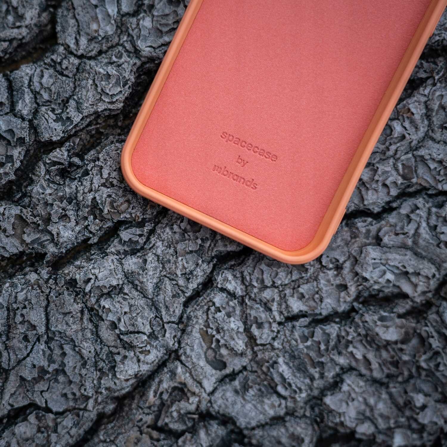 Pomarańczowe silikonowe etui z napisem \'spacecase by mbrands\' leży na tle szorstkiej, ciemnej kory drzewa.