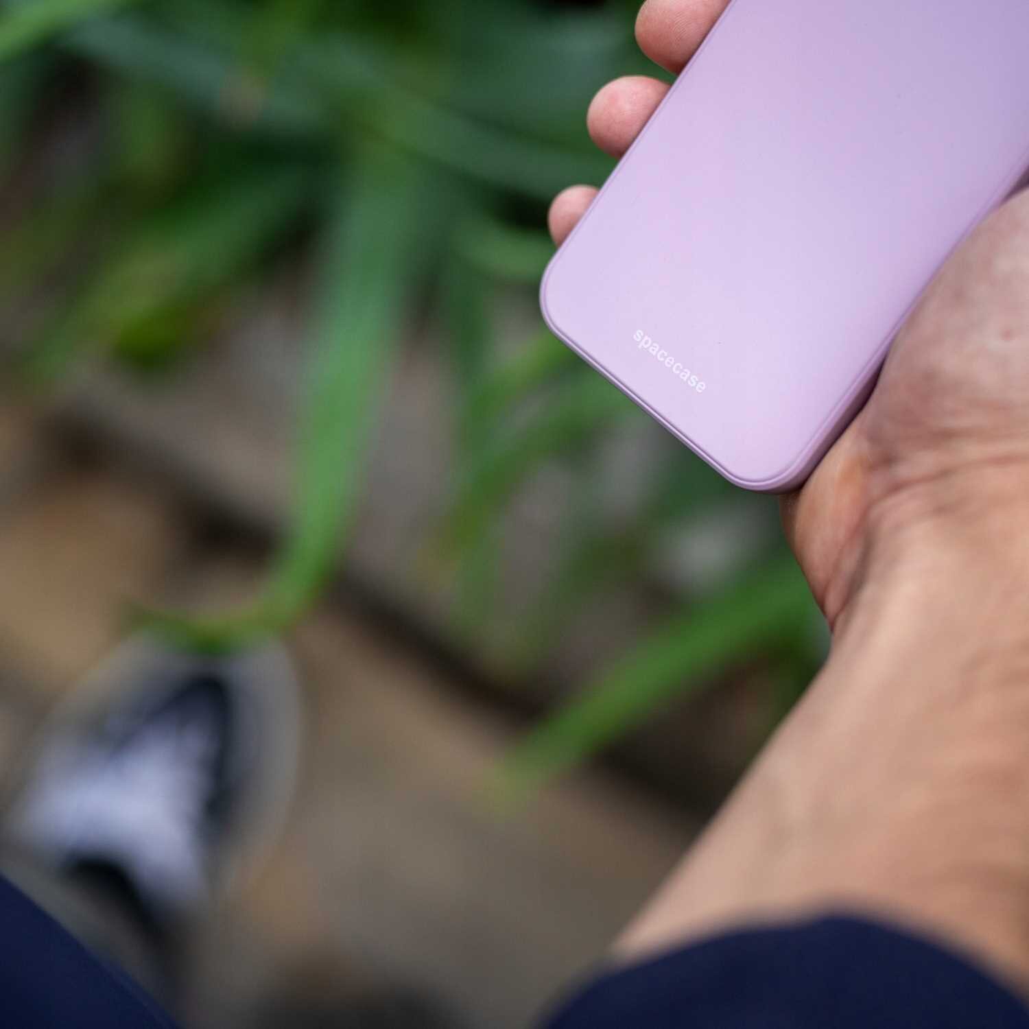 Osoba trzyma różowe etui na telefon z napisem \'spacecase\'. W tle widoczne są rozmyte zielone liście.