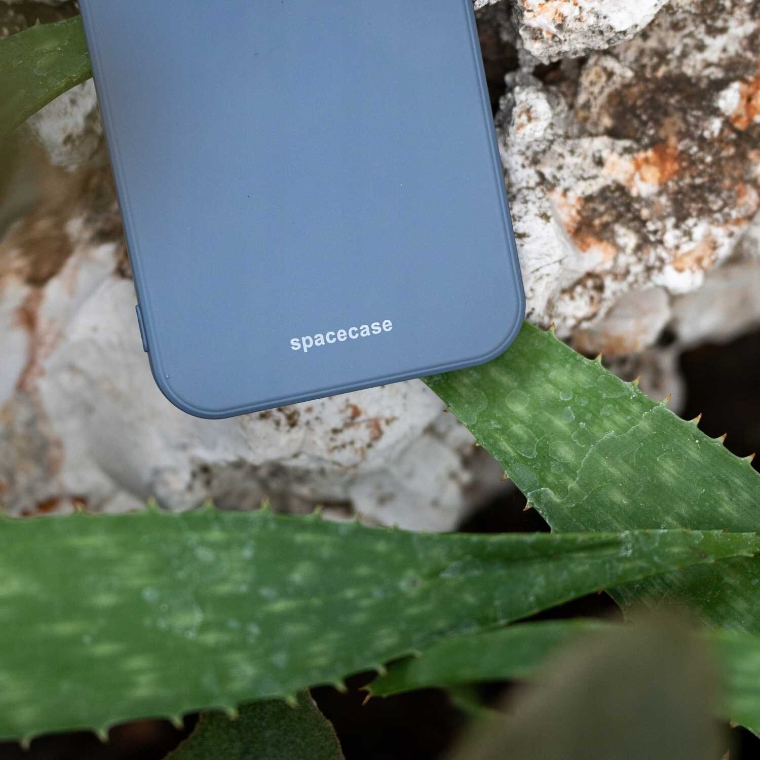Niebieskie silikonowe etui z napisem \'spacecase\' leży na tle kamieni i zielonych liści.