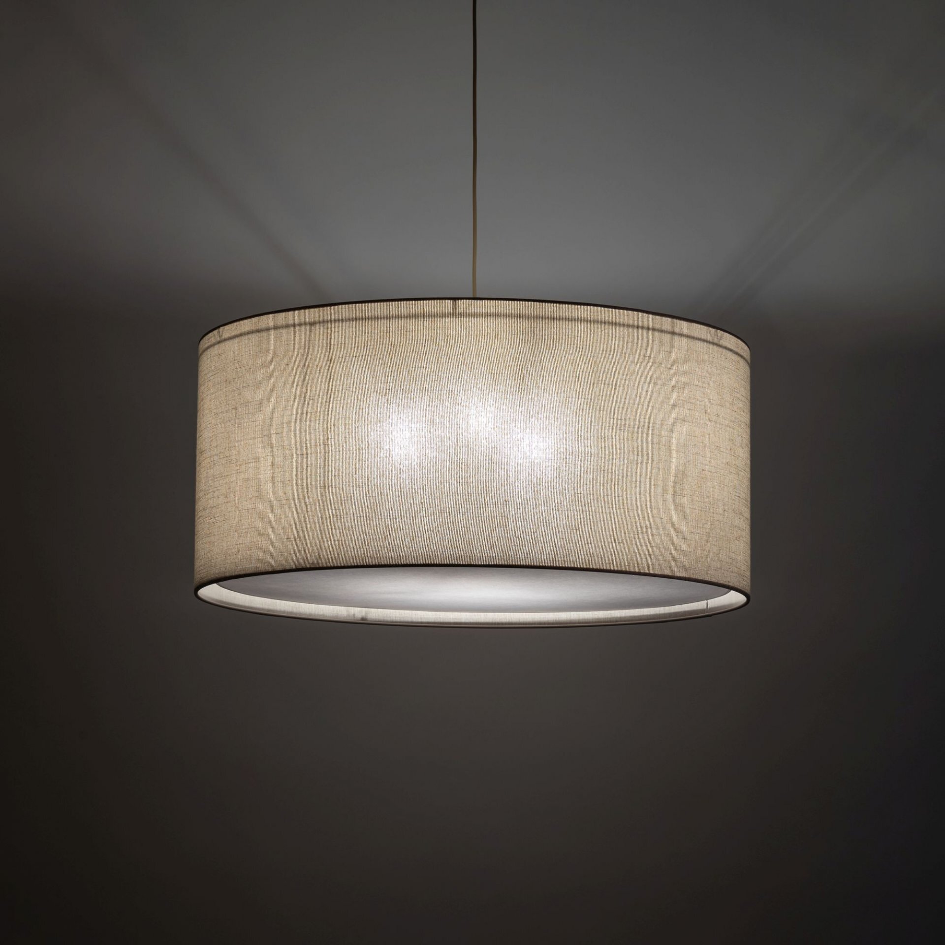 Na obrazie widoczna jest lampa wisząca z beżowym abażurem, emitująca miękkie światło. Lampa zawieszona jest na cienkim przewodzie, a tło jest ciemne.