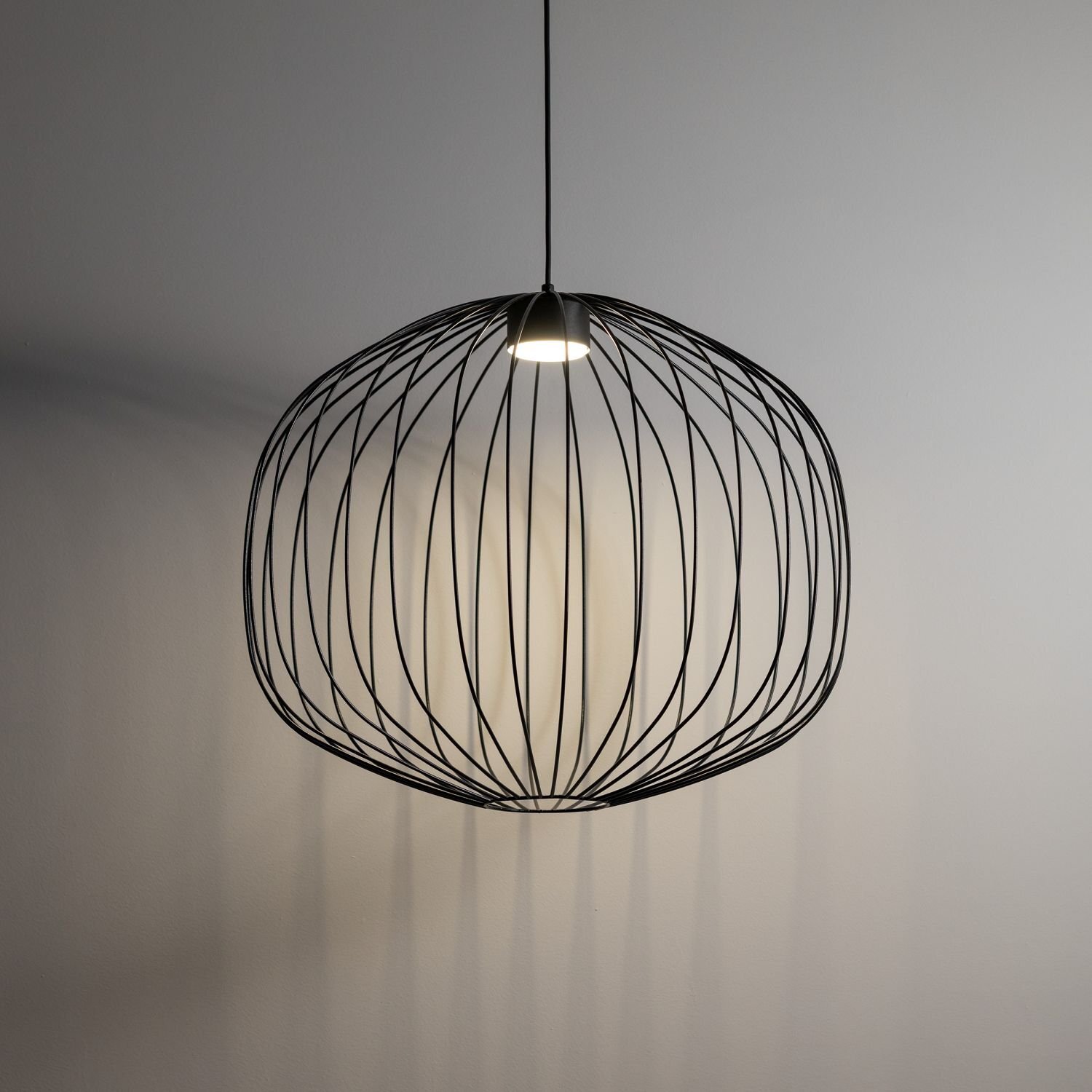 Czarna, wisząca lampa o kulistym kształcie z metalowych prętów, emitująca światło z centralnego punktu. Lampa zawieszona jest na cienkim kablu, a jej cień tworzy wzory na ścianie.