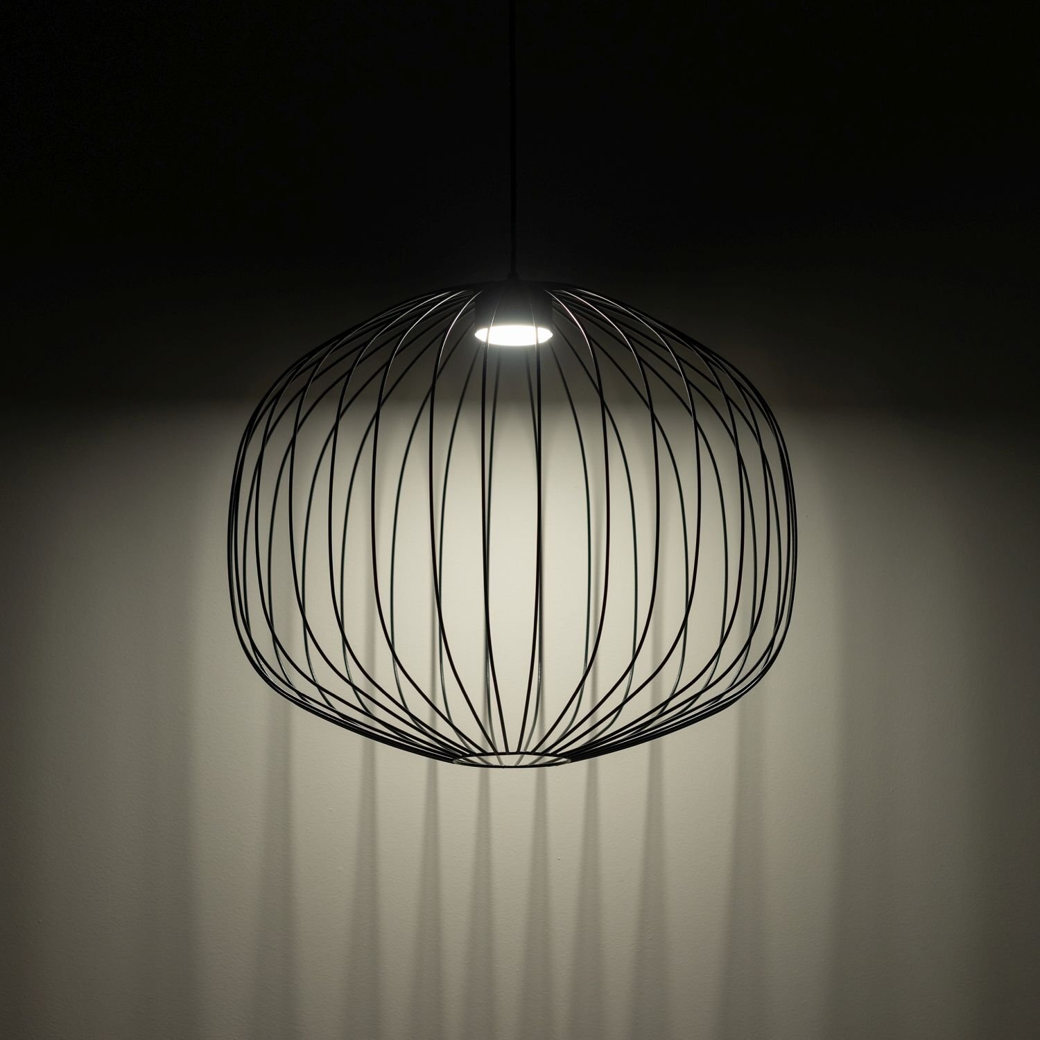 Czarna, wisząca lampa o kulistym kształcie z metalowych prętów, emitująca światło z centralnego punktu. Lampa tworzy cień na ścianie, podkreślając jej nowoczesny design.