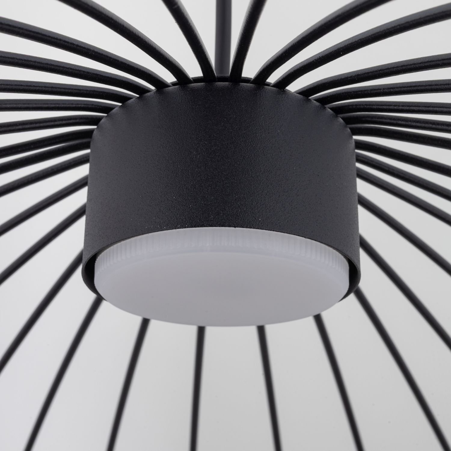 Zbliżenie na czarną, metalową lampę wiszącą o nowoczesnym designie z promieniście rozchodzącymi się prętami. Centralna część lampy jest cylindryczna z białym, matowym kloszem.